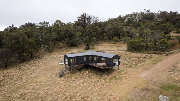 Eine moderne, dunkel gefärbte Hütte mit einer Terrasse, umgeben von trockenem Gras und Bäumen.