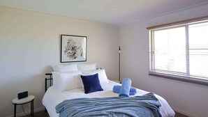 Ein Schlafzimmer mit Bett, Kissen, Decke, einem Nachttisch mit einem Gerät, einer Stehlampe und einem Fenster mit Jalousien.