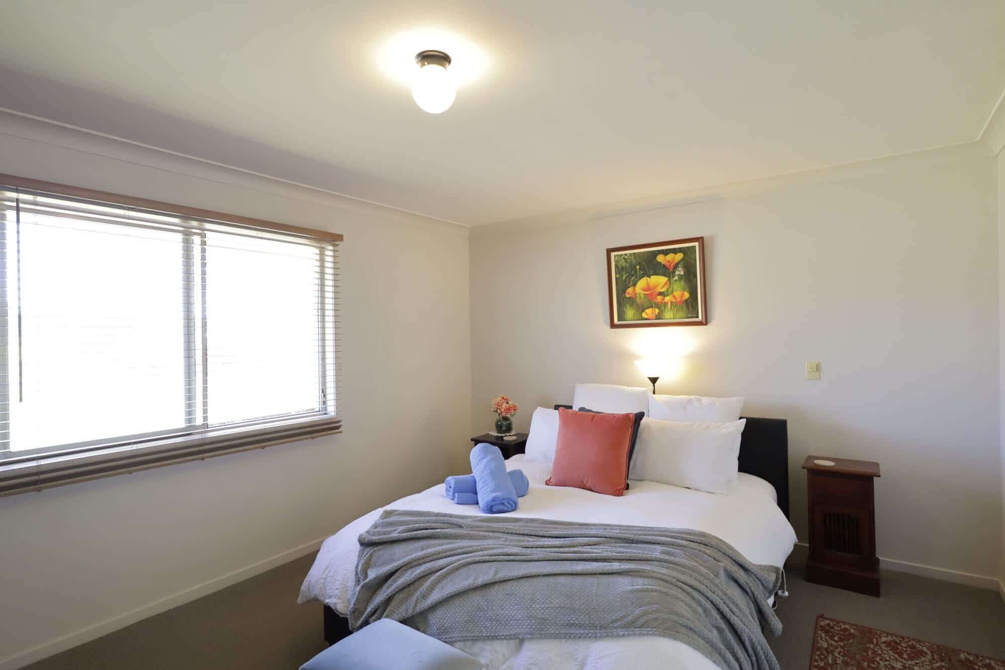 A bedroom with a bed, a nightstand, a lamp, a painting, and a window with blinds.