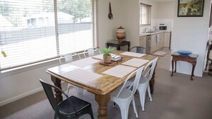 Ein Essbereich mit Holztisch und weißen Stühlen, im Hintergrund eine Küche und ein Fenster mit Jalousien.