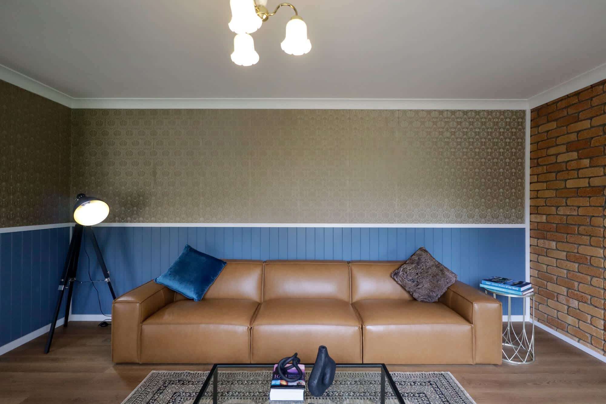 A living room with a brown leather sofa, a blue and brown patterned wall, a brick accent wall, a glass coffee table with books and a black object, and a floor lamp.
