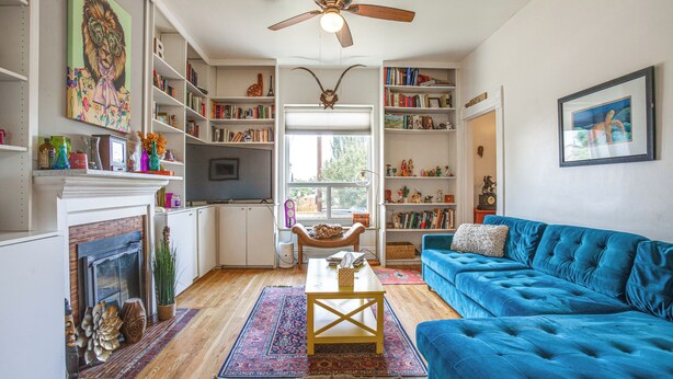 Una sala de estar con chimenea, un sofá azul, un ventilador de techo y una estantería llena de libros.