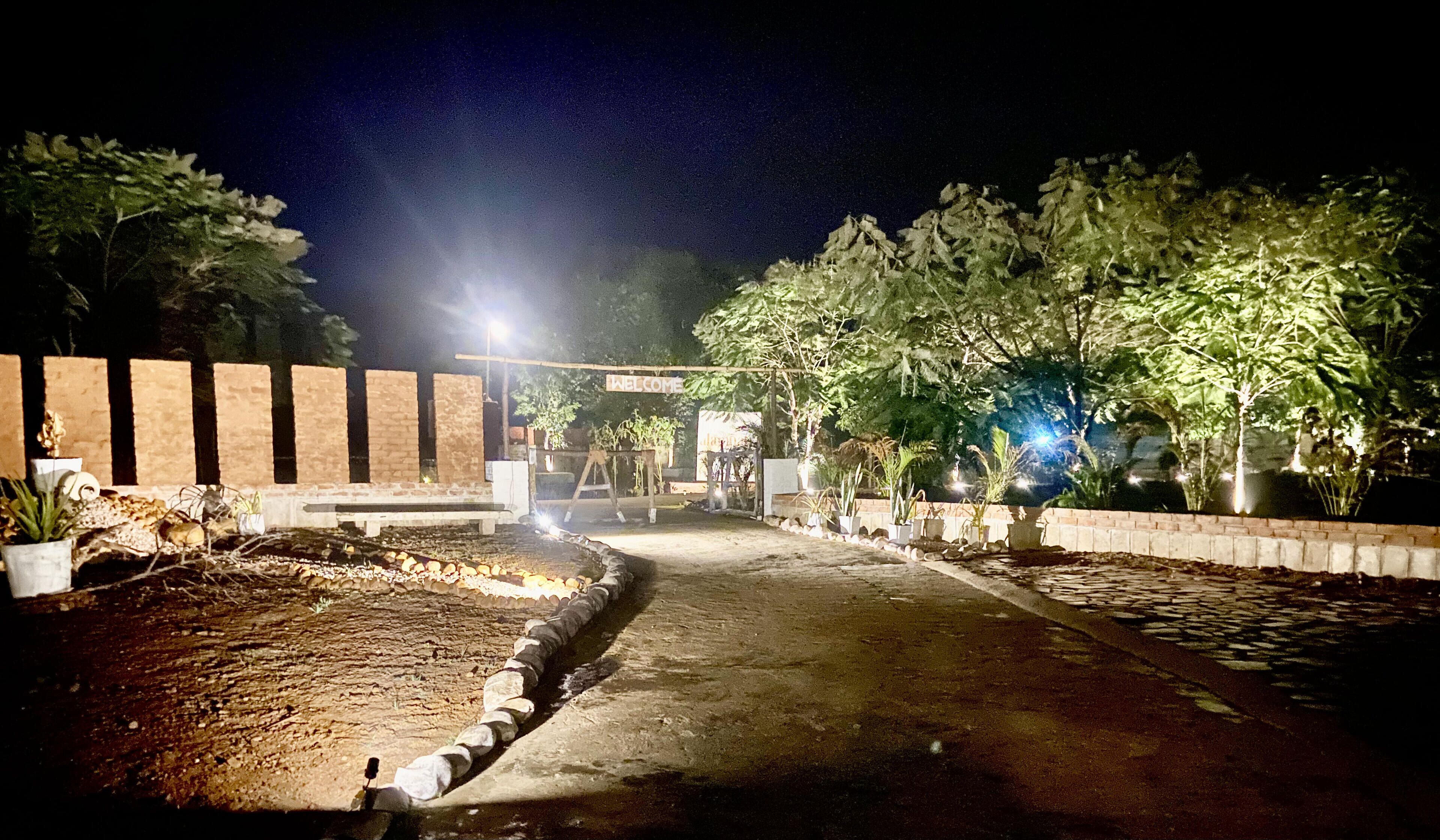 A nighttime view of a paved pathway leading to a building entrance with a 'WELCOME' sign, surrounded by potted plants and trees.