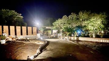 A nighttime view of a paved pathway leading to a building entrance with a 'WELCOME' sign, surrounded by potted plants and trees.