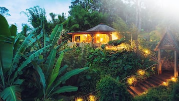 Una cabaña apartada ubicada en un exuberante jardín tropical, con una pequeña estructura parecida a un santuario cerca.