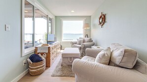 A living room with a sofa, ottoman, and a view of the sea through large windows. - Mtn-View Camano Island Retreat Overlooking Beach (Camano)