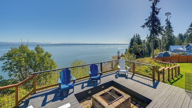 A deck with chairs overlooking a lake, a fire pit, and a view of a house and forest. - Mtn-View Camano Island Retreat Overlooking Beach (Camano)