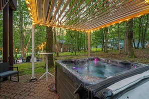 A hot tub under a pergola with string lights, surrounded by trees and a patio area with seating.