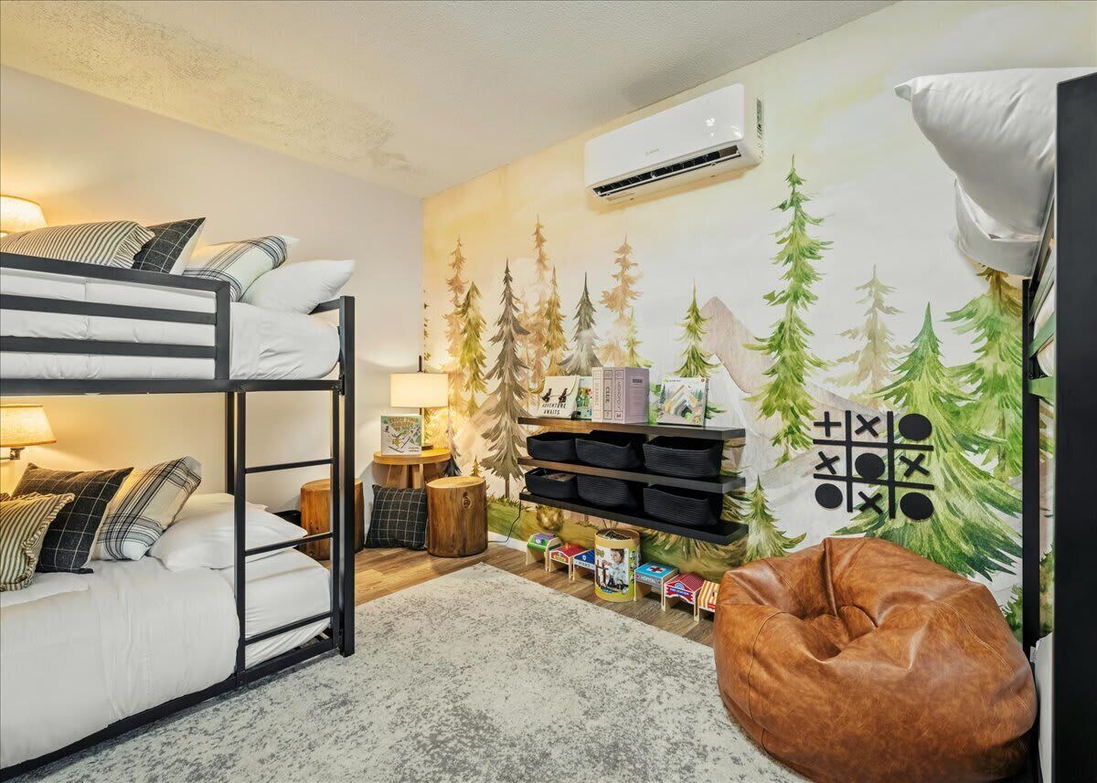 Una habitación acogedora con literas, un sillón orejero, aire acondicionado de pared y un estante con cajas de almacenamiento y un juego de tres en raya.