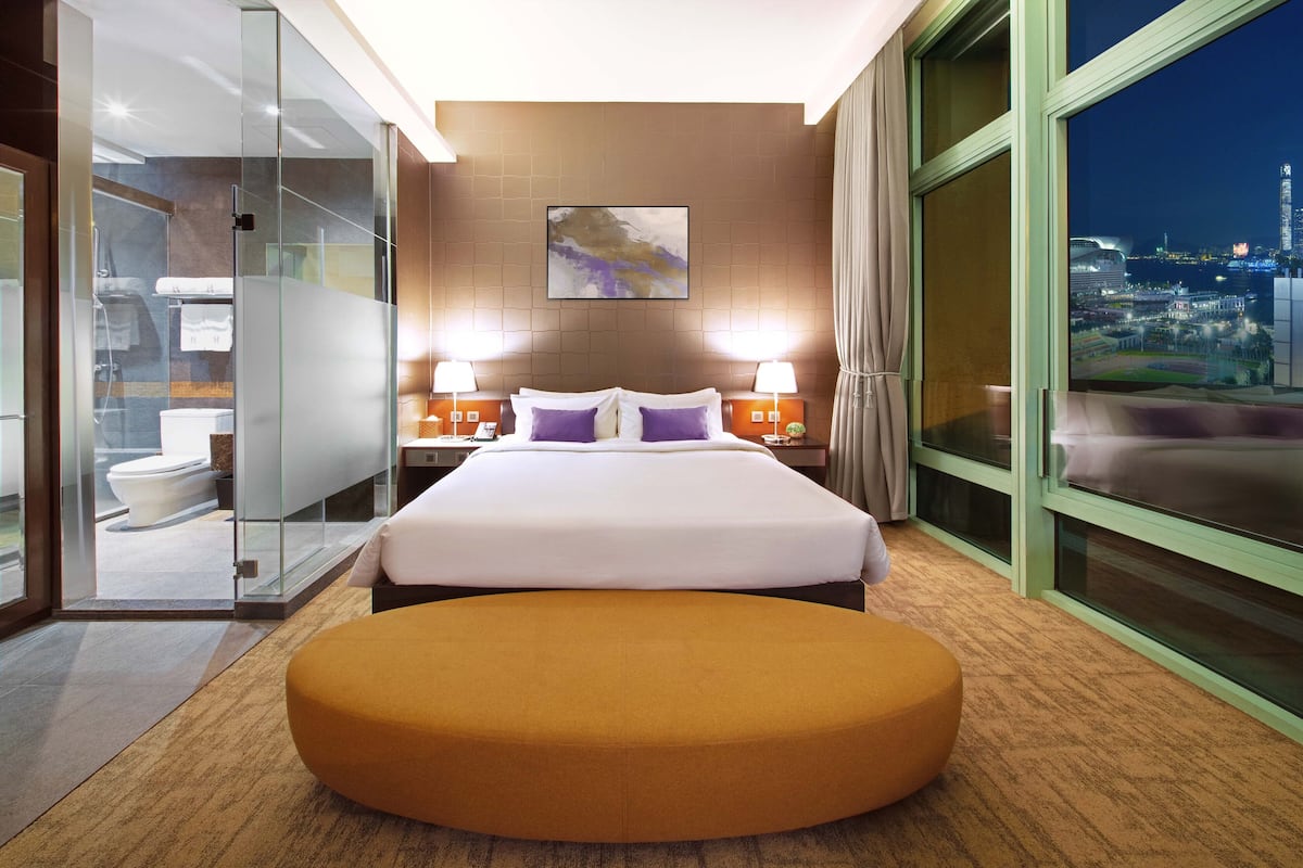 Quarto de hotel moderno com uma cama grande, box de vidro para o chuveiro e vista para a cidade.