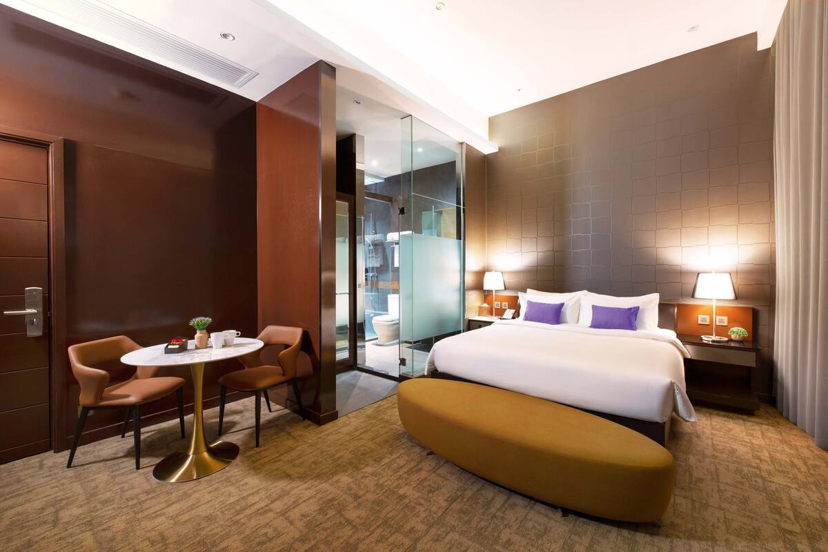 Quarto de hotel com uma cama, uma escrivaninha com cadeiras, uma mesinha com uma planta e um banheiro visível através de uma porta de vidro.