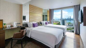 A hotel room with two beds, a desk, and a chair. There is a large window with a city view.