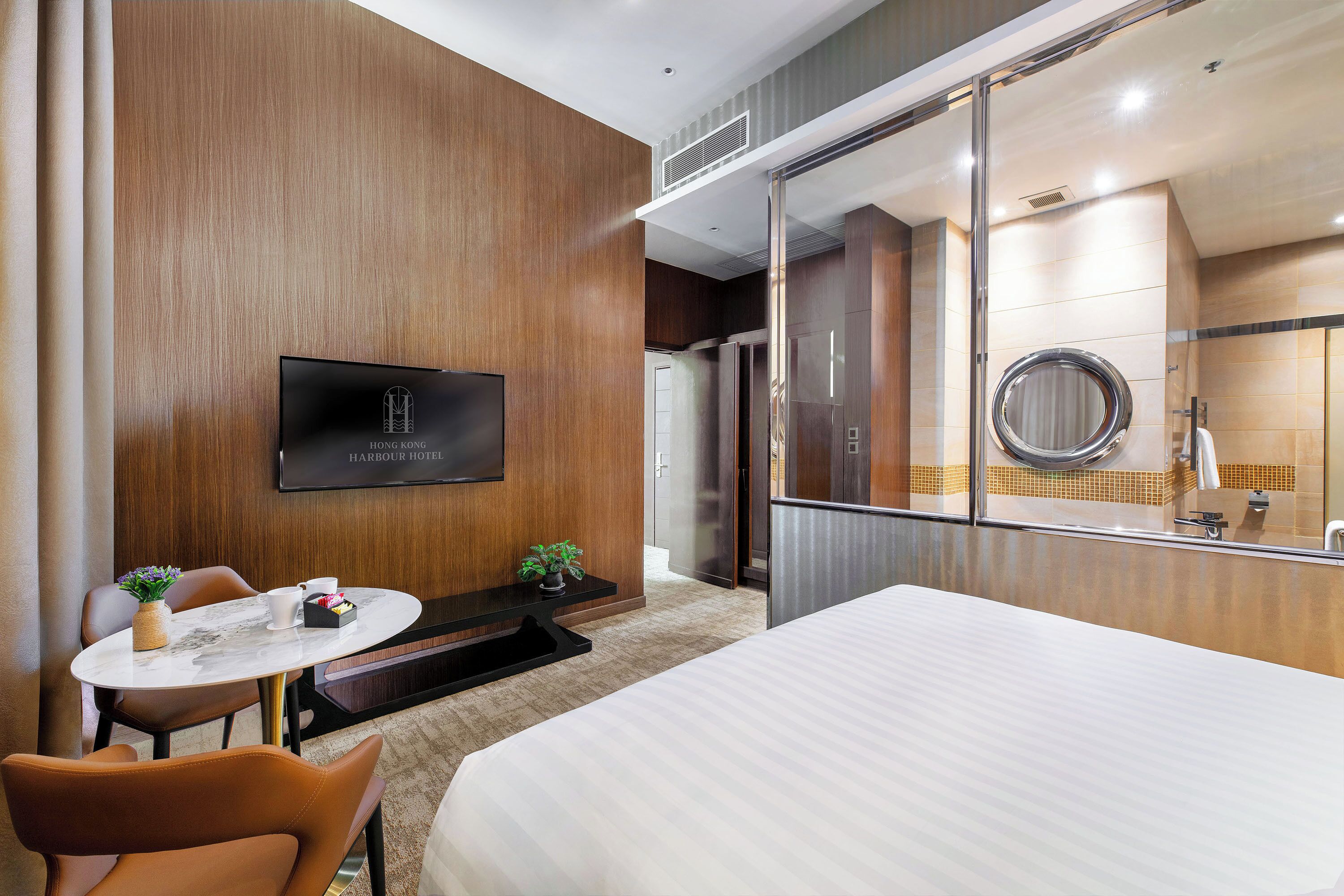 A hotel room with a bed, a desk with a chair, a television, and a bathroom visible through a glass partition.