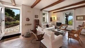 A spacious living room with a white sofa, wicker chairs, and a fireplace. There is a stone pathway outside and a door leading to a garden.