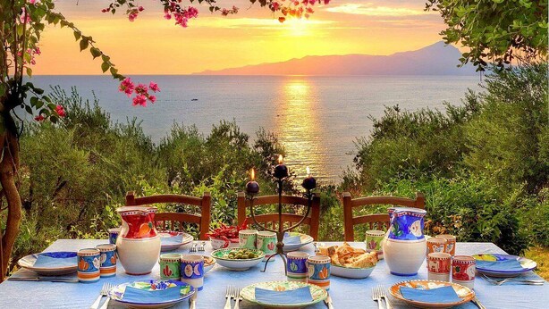 A table set for a meal with a view of the sea and mountains at sunset.