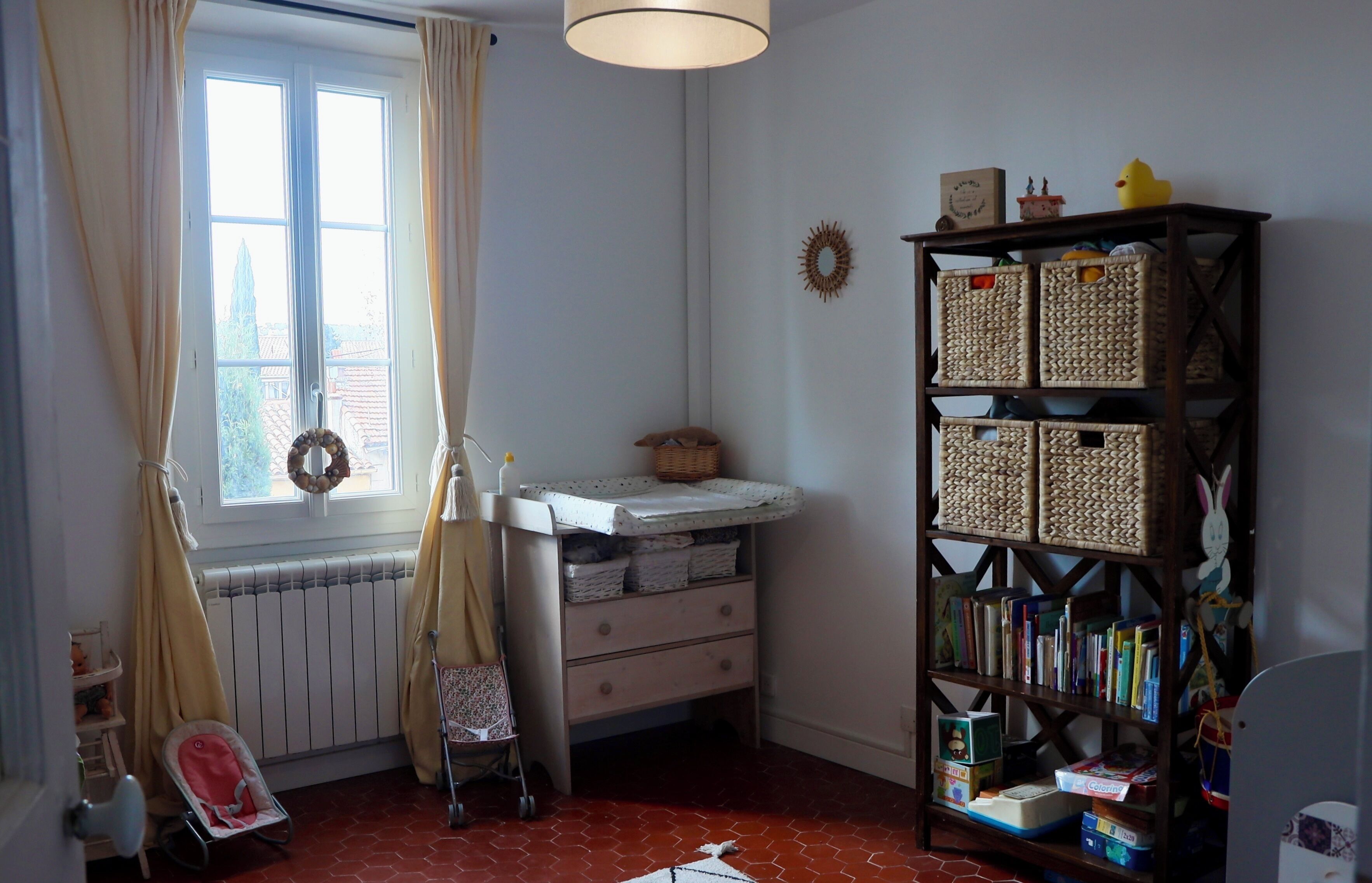 Une pièce avec un radiateur, un transat pour bébé, une bibliothèque et une fenêtre avec des rideaux.