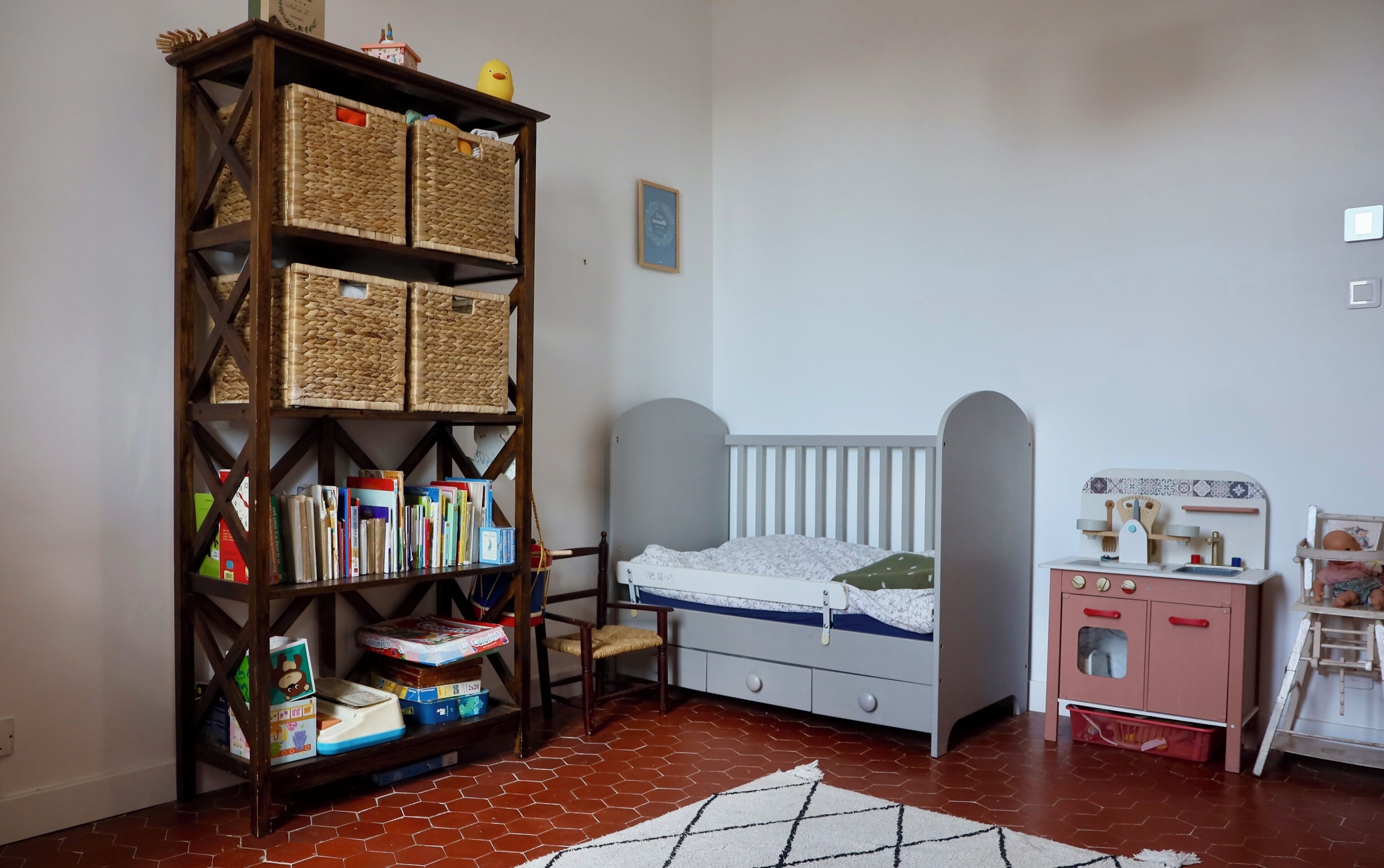 A child's room with a crib, a bookshelf, a small dresser, and a toy kitchen set.