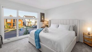 A modern hotel room with a large window, a white bed, a bedside table with a lamp, and a view of the street and other buildings.
