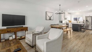 A modern living room with a flat-screen TV, white armchairs, a wooden coffee table, and a dining area with a small island.