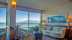 A spacious living area with a large window offering a view of the ocean and mountains. There is a sofa, a coffee table with a plant, and a painting on the wall. - Royal Kahana 404 (Lahaina)
