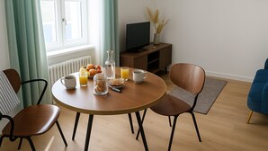 Ein Essbereich mit einem runden Tisch, Stühlen und Frühstücksangeboten. Im Hintergrund steht ein Fernseher auf einem Beistelltisch.