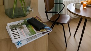 Ein Zimmer mit Holzboden, einer Glaskaraffe mit weißen Blumen, einem Stuhl, einem Tisch mit Tasse und einer Auswahl an Croissants sowie einem Fenster mit Vorhängen.