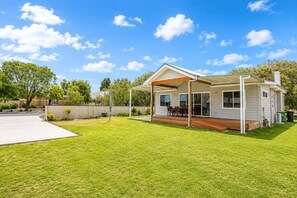 Ein eingeschossiges Haus mit überdachter Terrasse, Rasen und Holzdeck.