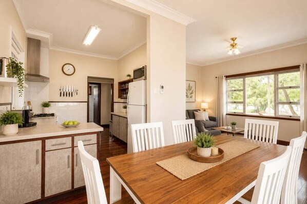 A modern kitchen with white cabinets, a wooden dining table, and a view of the outdoors through large windows.