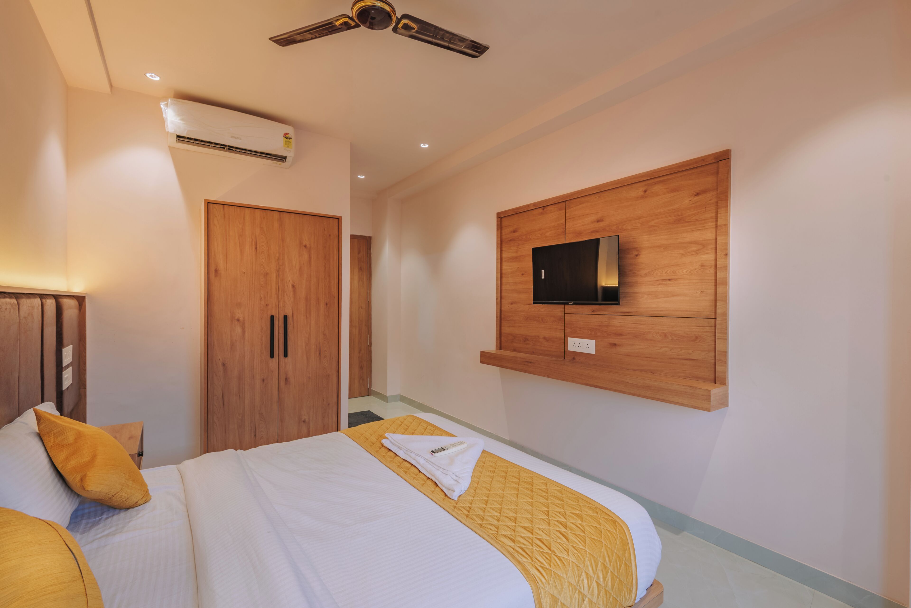 A hotel room with a bed, a wooden wardrobe, a mounted TV, and an air conditioning unit.