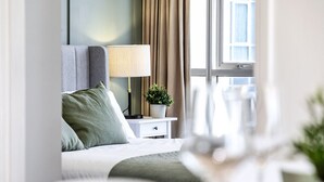 A modern hotel room with a neatly made bed, a bedside lamp, a small potted plant, and a window with curtains.