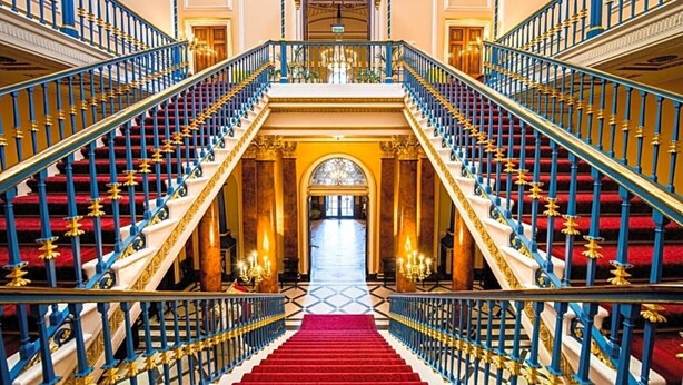 Una imponente escalera con alfombra roja, barandillas azules y doradas, y un candelabro en la parte superior.