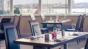 Un comedor con mesas dispuestas para dos, sillas y vistas a un paisaje urbano a través de amplias ventanas.