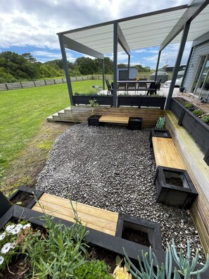 Un área de estar al aire libre techada, con bancos de madera y jardineras, un camino de grava y vistas a un campo cubierto de césped y árboles al fondo.