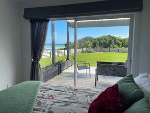 A bedroom with a bed, a green throw, a floral bedspread, and a view of a grassy yard and ocean through a sliding glass door. - The ‘Blue Bach’. Family memories waiting to be made. (Ōpunake)