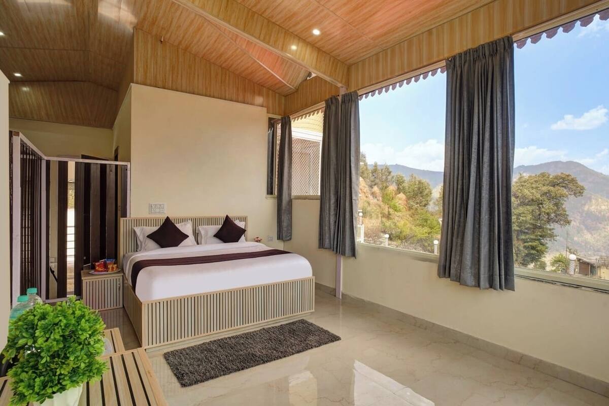 A bedroom with a bed, a small table, a potted plant, and a view of mountains through large windows.