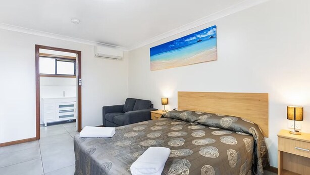 A hotel room with a bed, a nightstand, a lamp, a painting of a beach, a sofa, and a wardrobe. - Palm Valley Motel (Tarro)