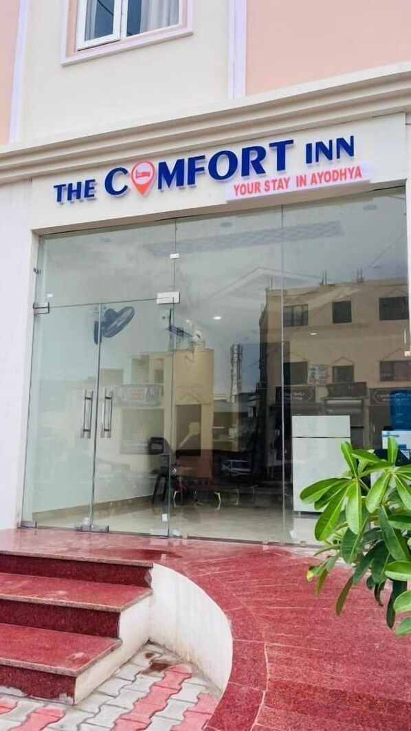 The exterior of The Comfort Inn with a clear glass facade, a red-tiled entrance, and a sign indicating the location in Ayodhya. - The Comfort Inn (Faizabad)