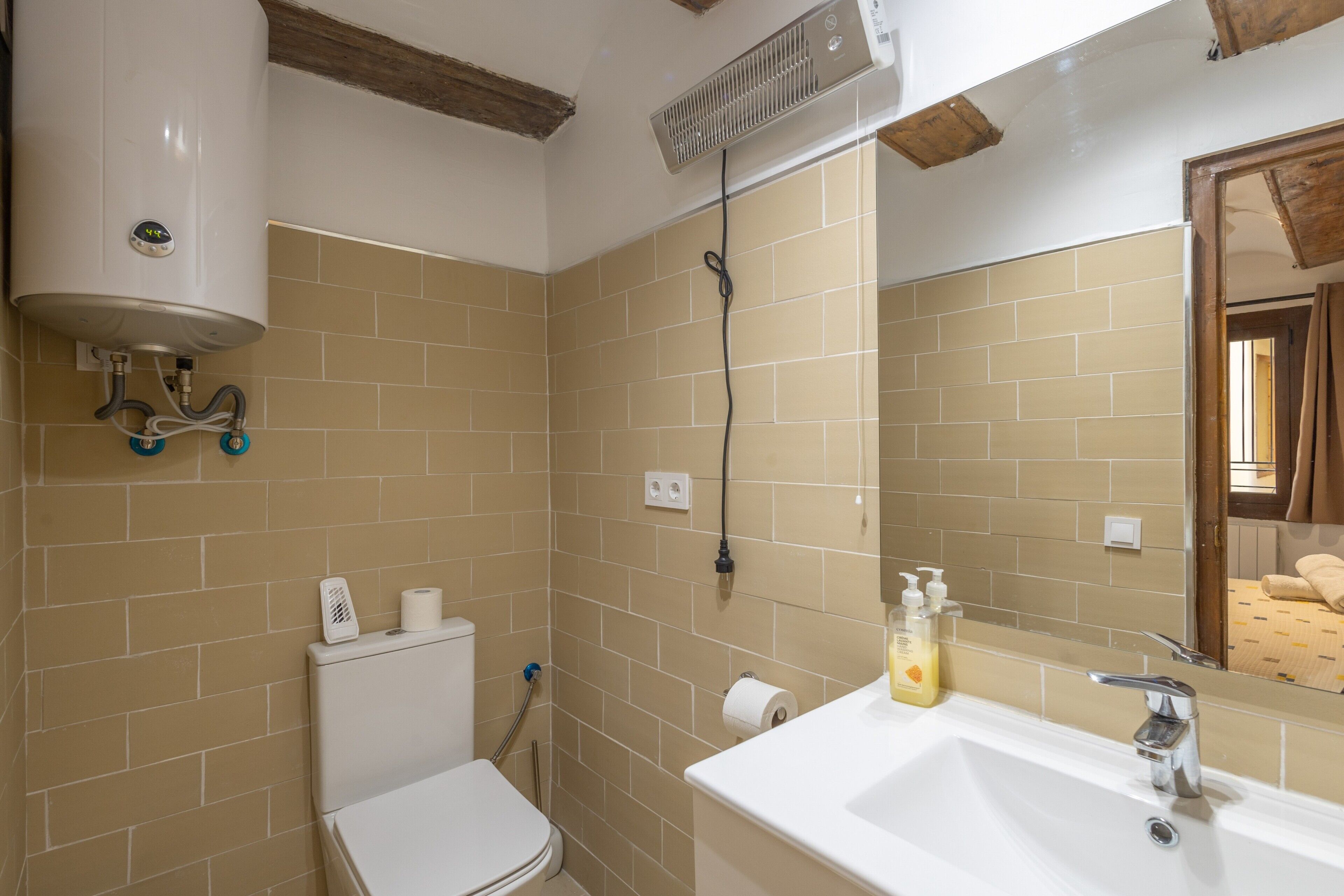 Une salle de bain avec des toilettes blanches, des murs recouverts de carreaux beiges, un lavabo blanc avec un miroir et un radiateur blanc fixé au mur.