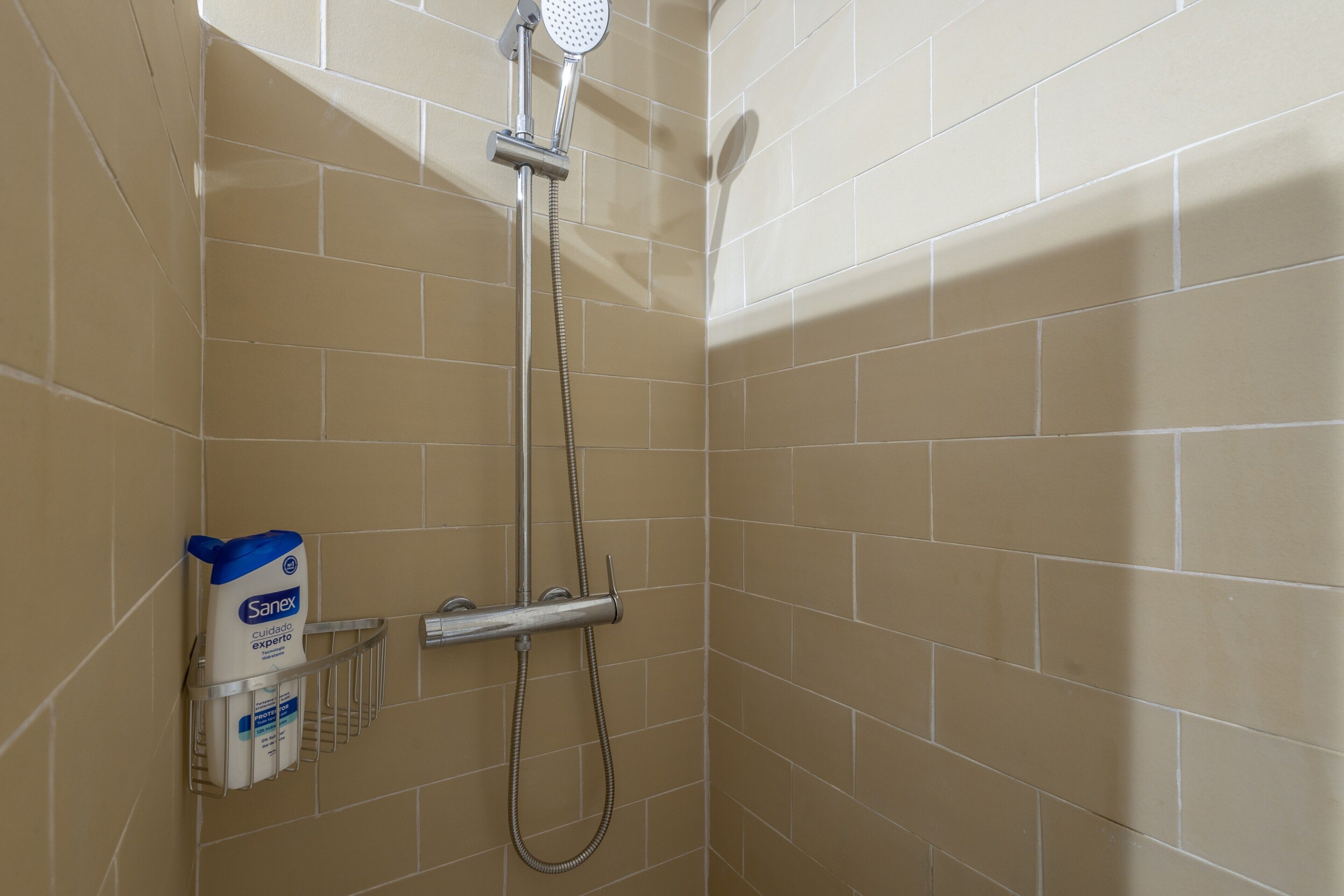 Ducha con azulejos color beige, cabezal de ducha de mano y dispensador de jabón en la pared.
