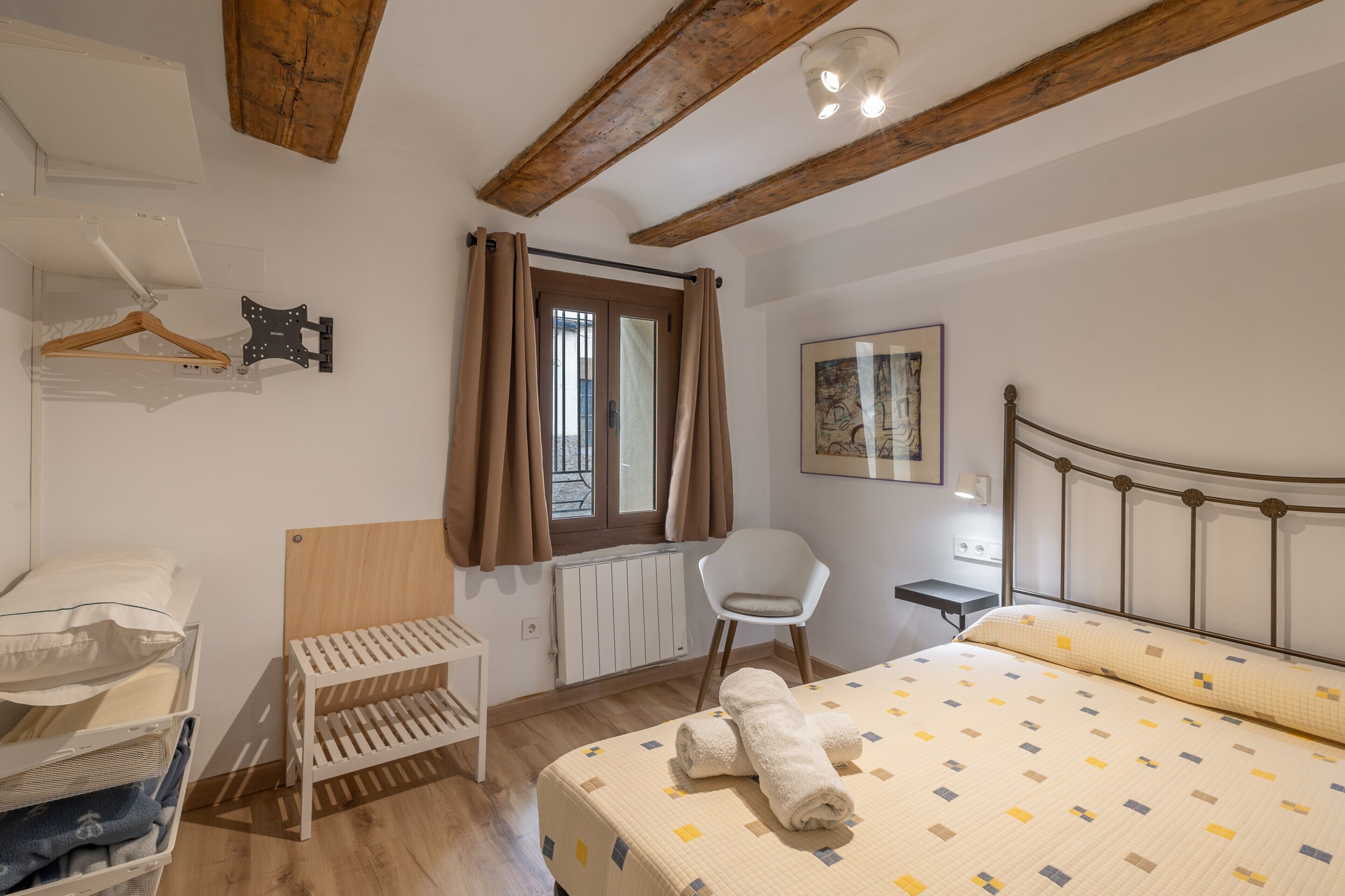 A bedroom with a bed, a chair, a radiator, a window with curtains, a wooden ceiling, and a wall-mounted hanger.