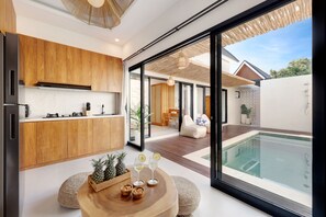 A modern kitchen with wooden cabinets, a dining area with a round wooden table, and a view of a swimming pool through large glass doors. - Villa Summer Cinta (Seminyak)