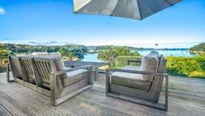 A deck with two wooden lounge chairs and a large umbrella, overlooking a scenic view of a lake and hills. - Shelley Beach Homestead (Waiheke Island)