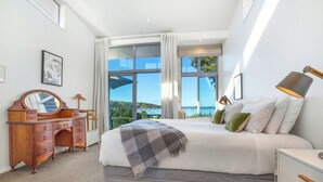 A bedroom with a large bed, a wooden dresser, a mirror, and a view of the outdoors. - Shelley Beach Homestead (Waiheke Island)