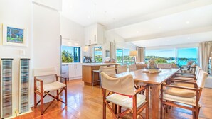A spacious dining area with a long wooden table and multiple chairs, a kitchen area with modern appliances, and large windows offering a view of the landscape. - Shelley Beach Homestead (Waiheke Island)