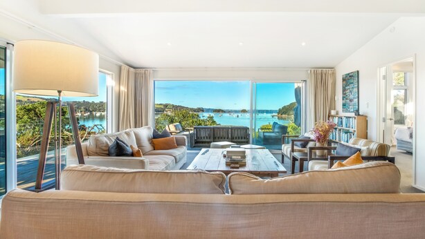 A spacious living room with a large window offering a scenic view of the sea and boats. There is a sofa set, a coffee table, and a bookshelf. - Shelley Beach Homestead (Waiheke Island)