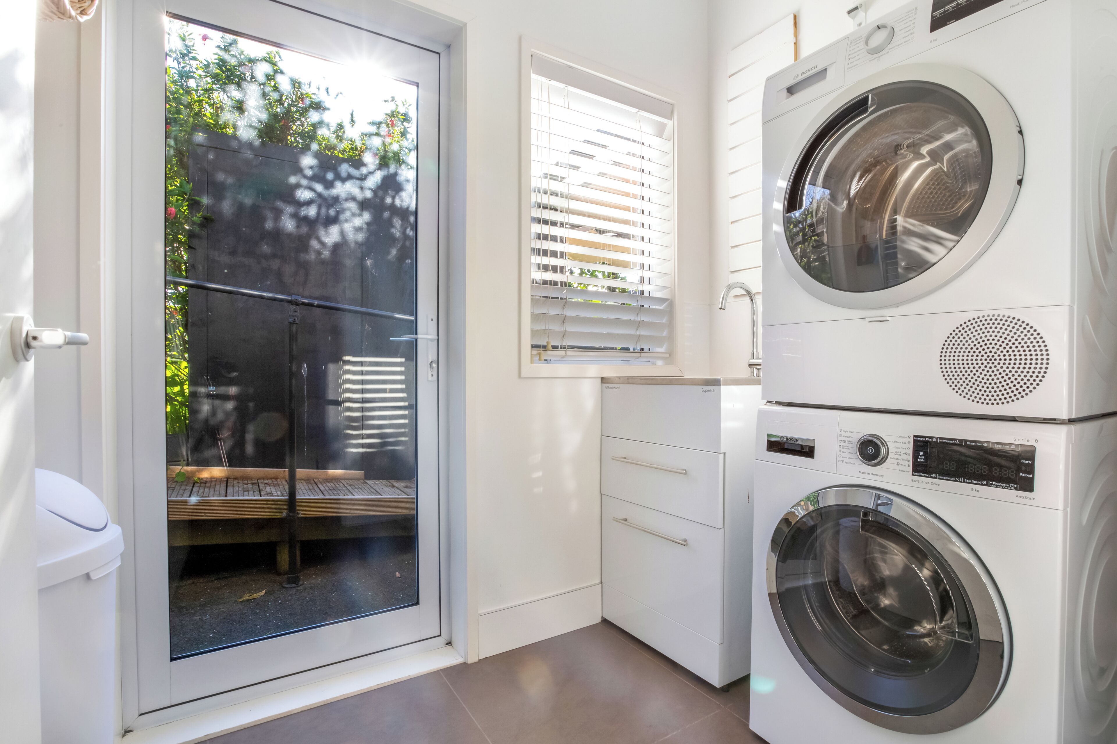 A laundry room with two stacked front-loading washing machines, a window with blinds, and a door leading to an outdoor area.