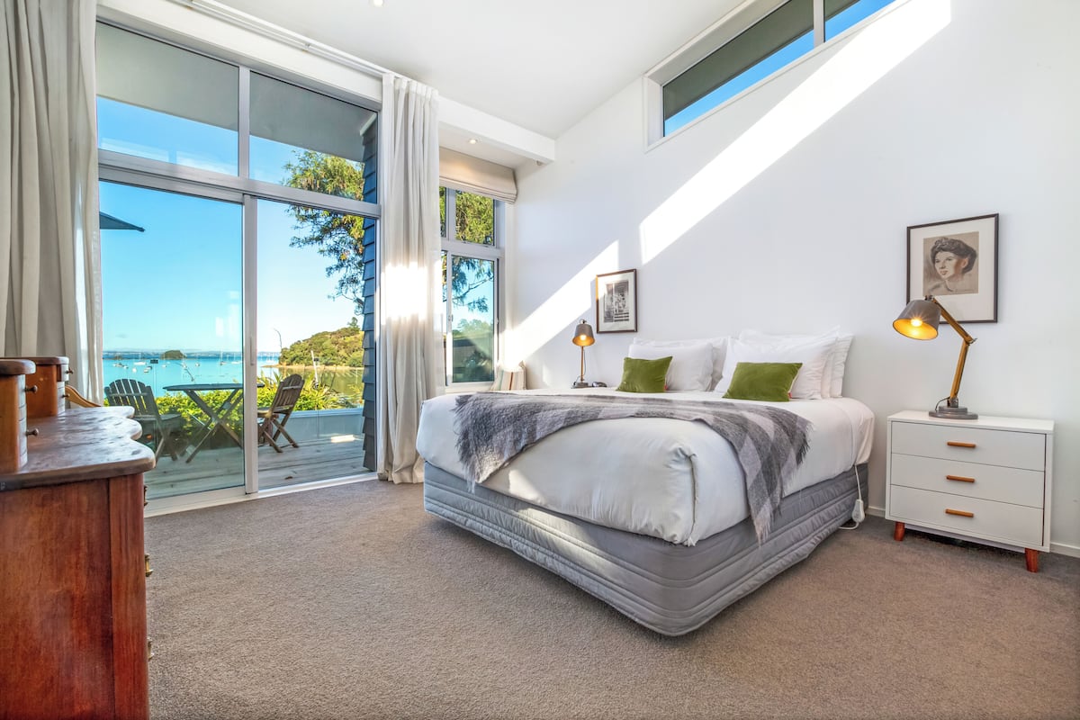 A bedroom with a large bed, a nightstand, a lamp, and a view of the outdoors.