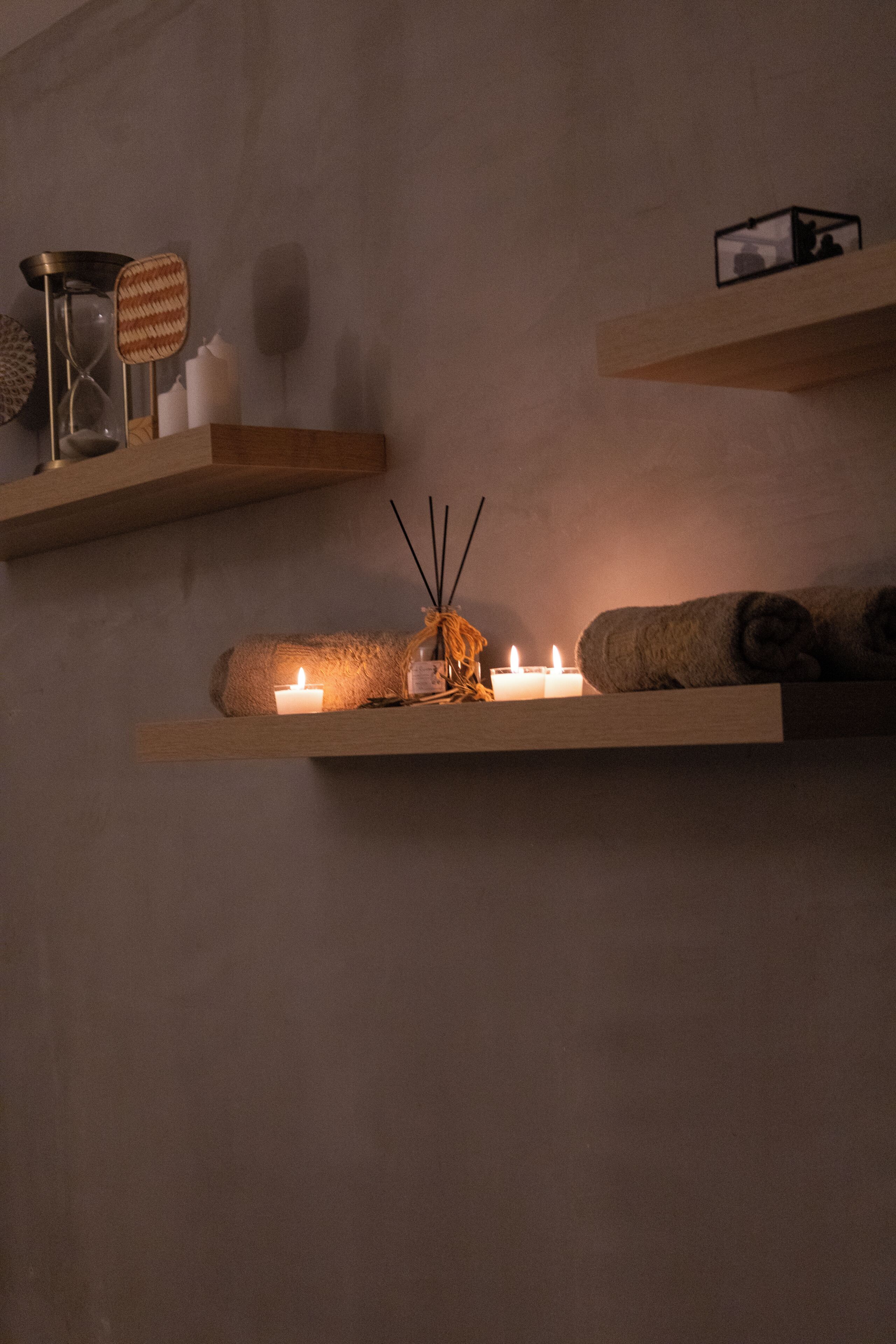 Uma parede com três prateleiras de madeira, cada uma com velas, um castiçal pequeno e uma toalha enrolada.