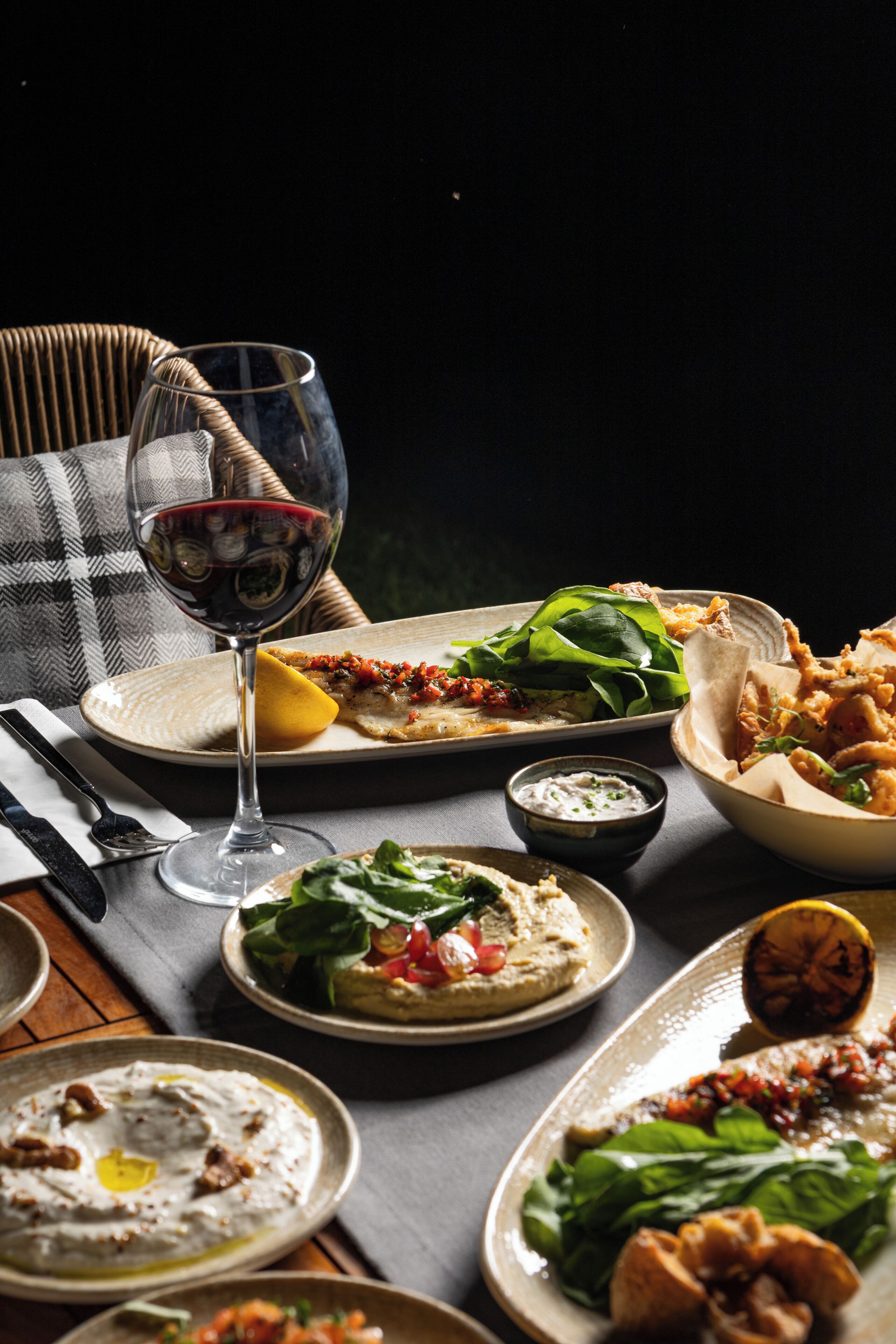 Una mesa con varios platos, una copa de vino tinto y una silla de mimbre.