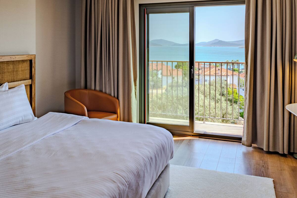 Ein Hotelzimmer mit Bett, Stuhl, Deckenventilator, Blick durch eine Schiebetür und einem Balkon.